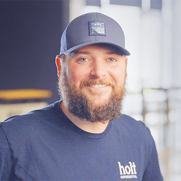 Male smiling worker with beard wearing Holt Experiential cap in modern industrial setting.