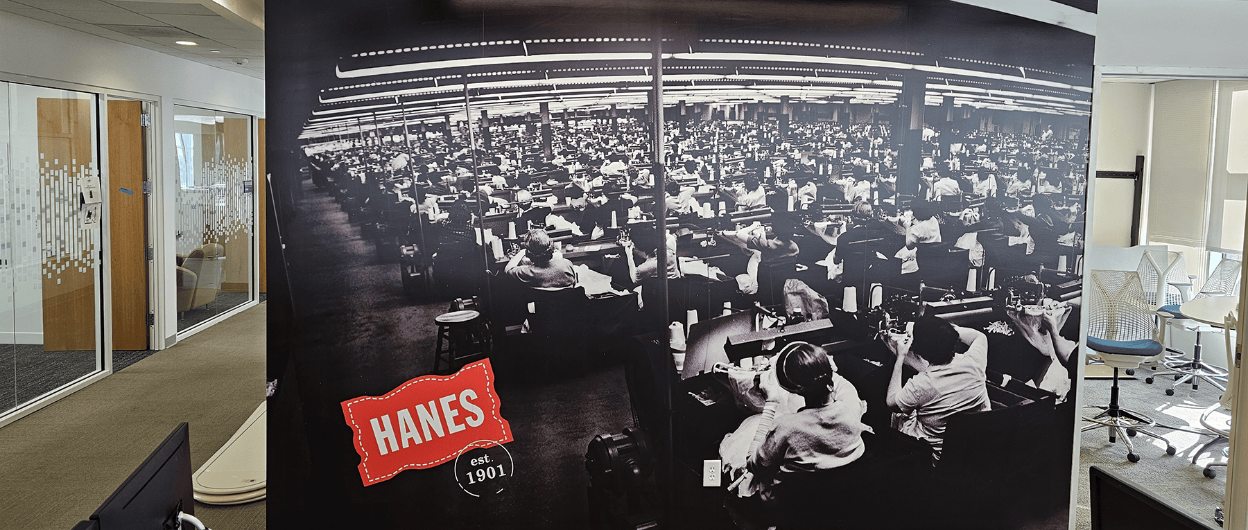 Office hallway at HanesBrands Park Building featuring a historic mural of a 1901 Hanes workshop. The artwork celebrates the company&rsquo;s heritage and provides context for its long-standing tradition of quality and innovation.