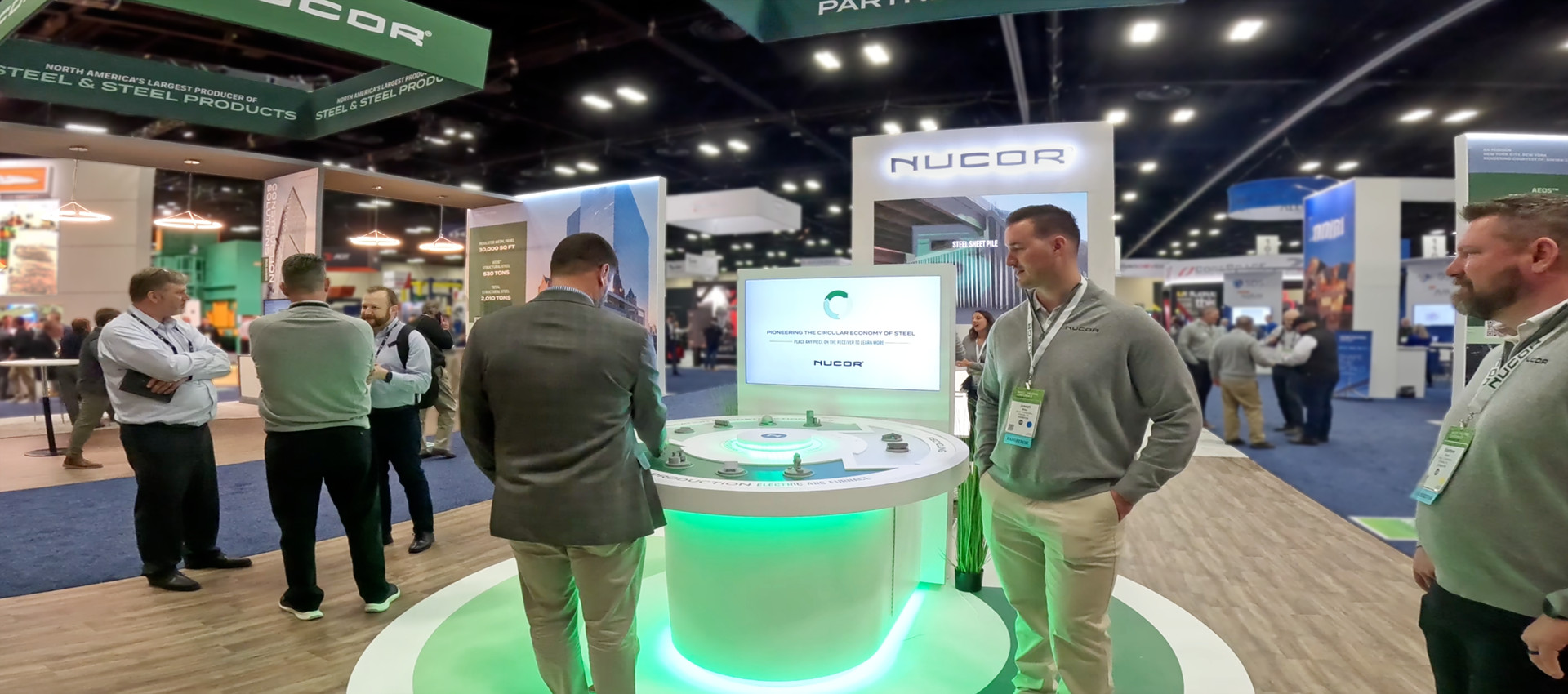 NUCOR sales team engaging customers at a trade show using a digital interactive table with a large video screen inside a custom exhibit