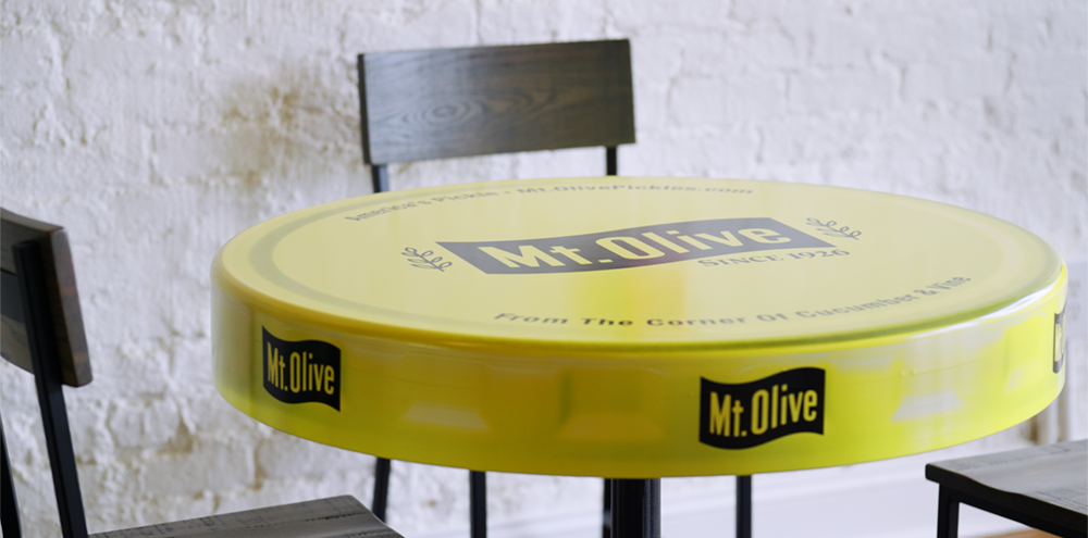 Round Mount Olive jar lid tables inside the Pickle Parlor where guests sit eating pickles and enjoying drinks.